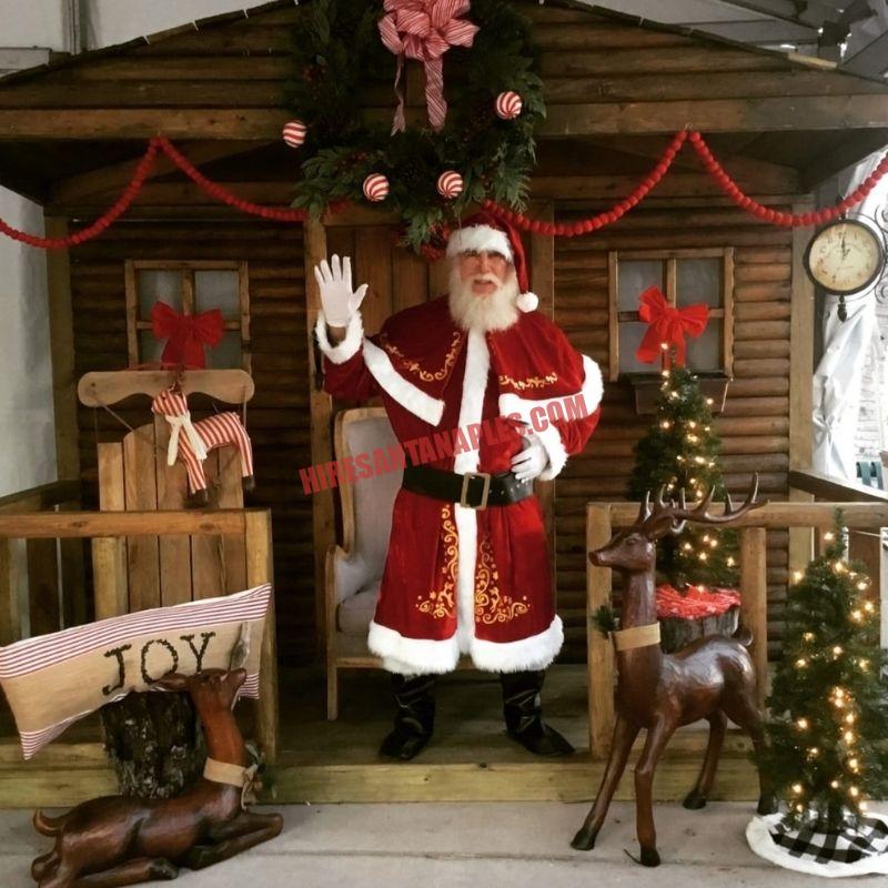 Real bearded Santa for hire in Naples, FL waving from a log-cabin porch beside a carved reindeer.