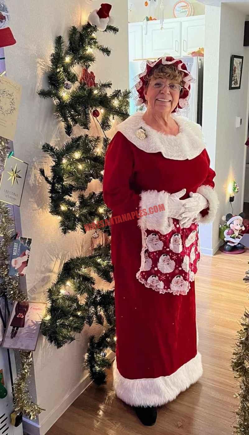 Mrs. Claus for hire in Naples, FL standing in a hallway by a slim lit tree in a red dress.