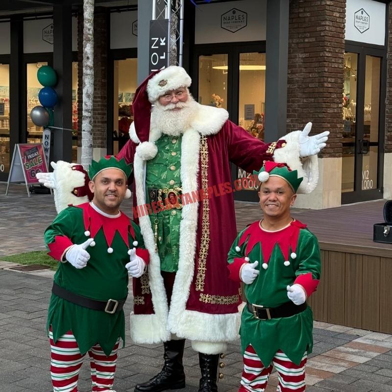 Santa Claus impersonator for hire in Naples, FL standing outdoors with two elves, thumbs up.