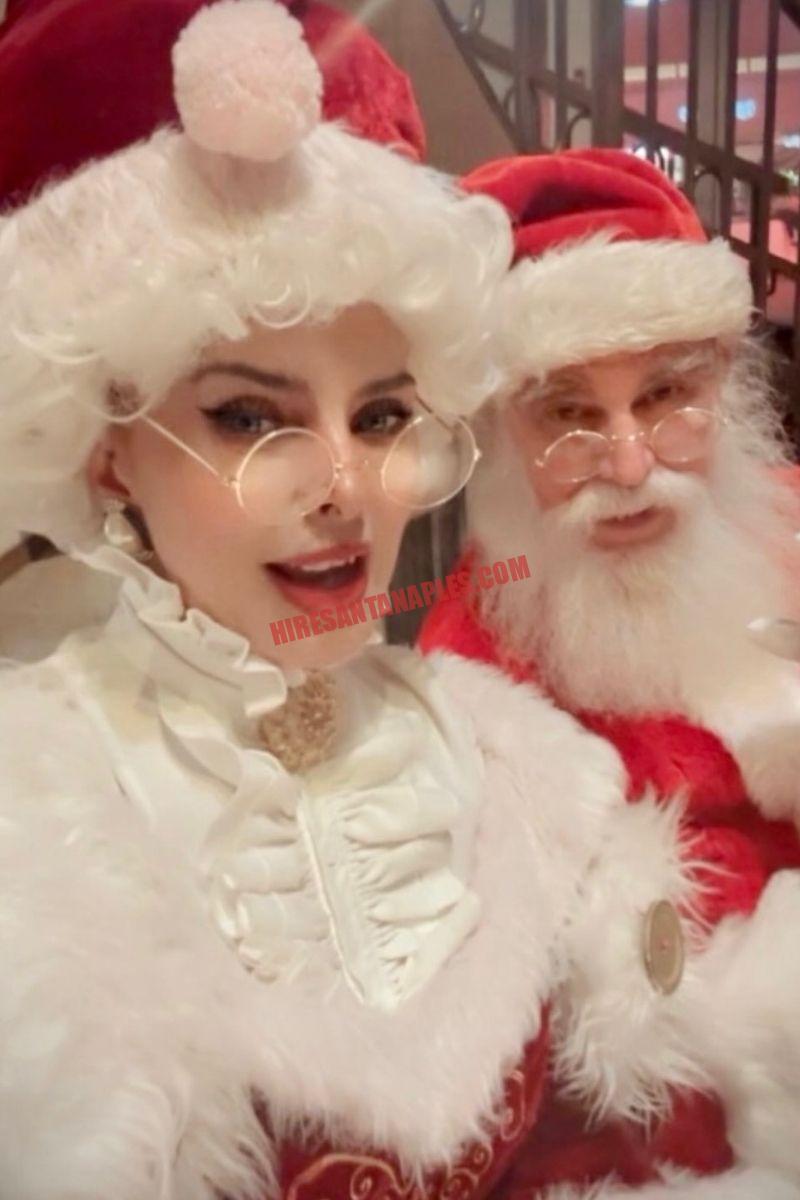 About Hire Santa Naples: Mrs. Claus in white ruffles standing beside Santa in a red suit.